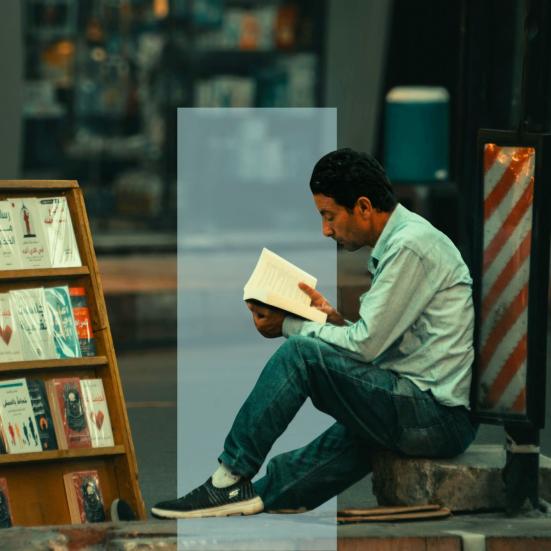 En mand sidder på en kantsten og læser i en bog ved siden af et bogstativ.